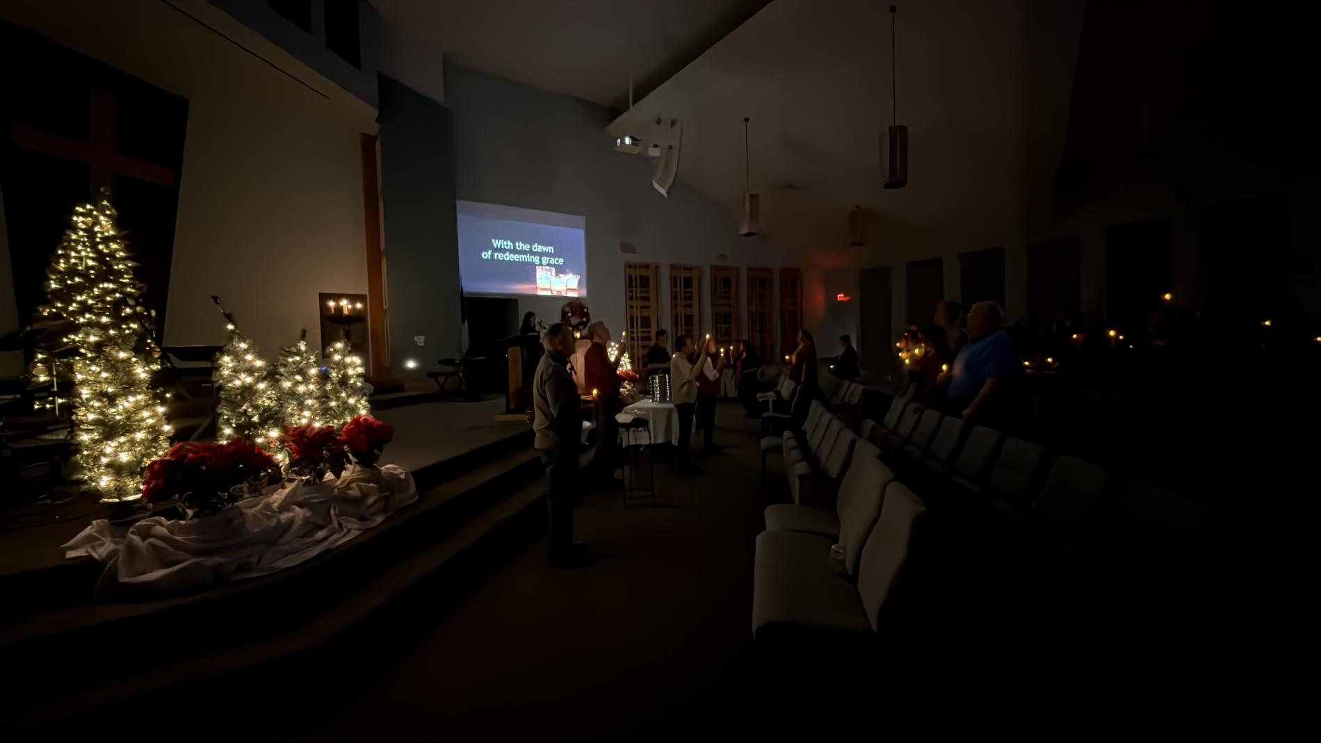 Dimly lit church interior with Christmas trees and a small group gathered.