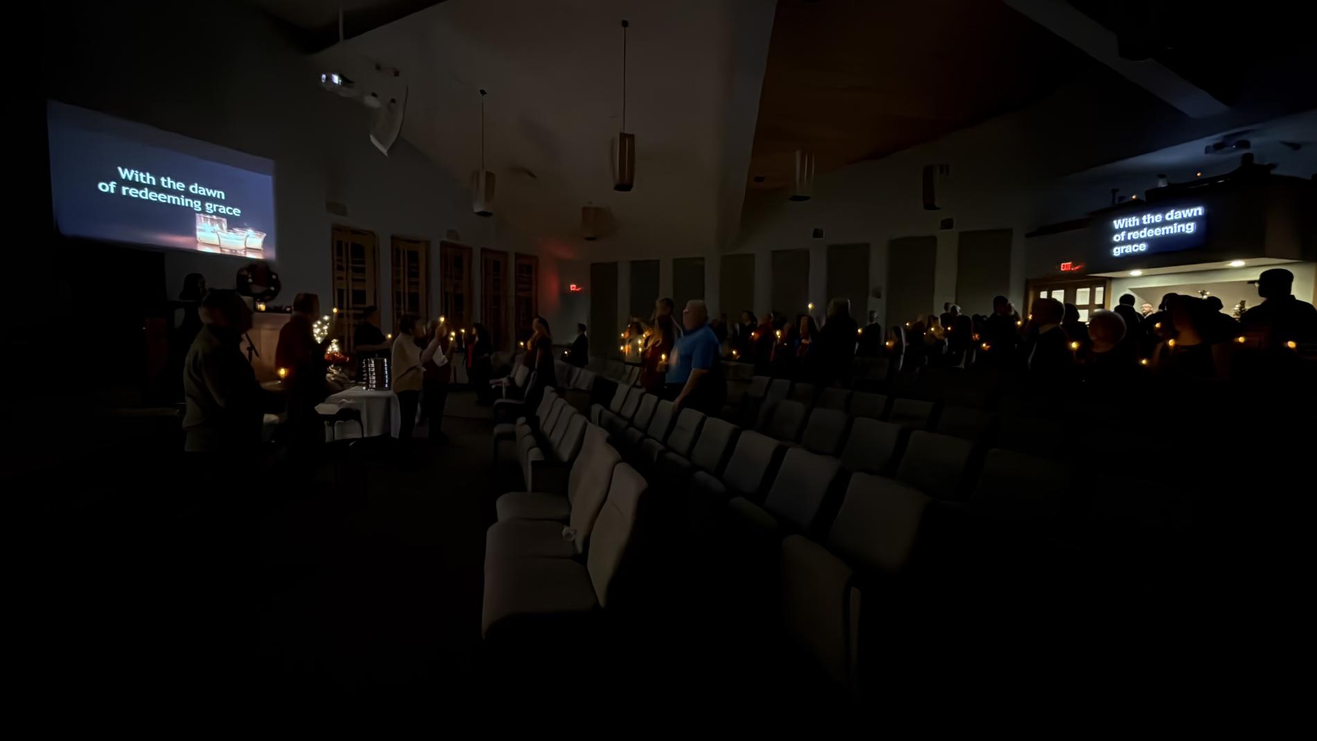 Dimly lit hall with people holding candles, seated in rows.