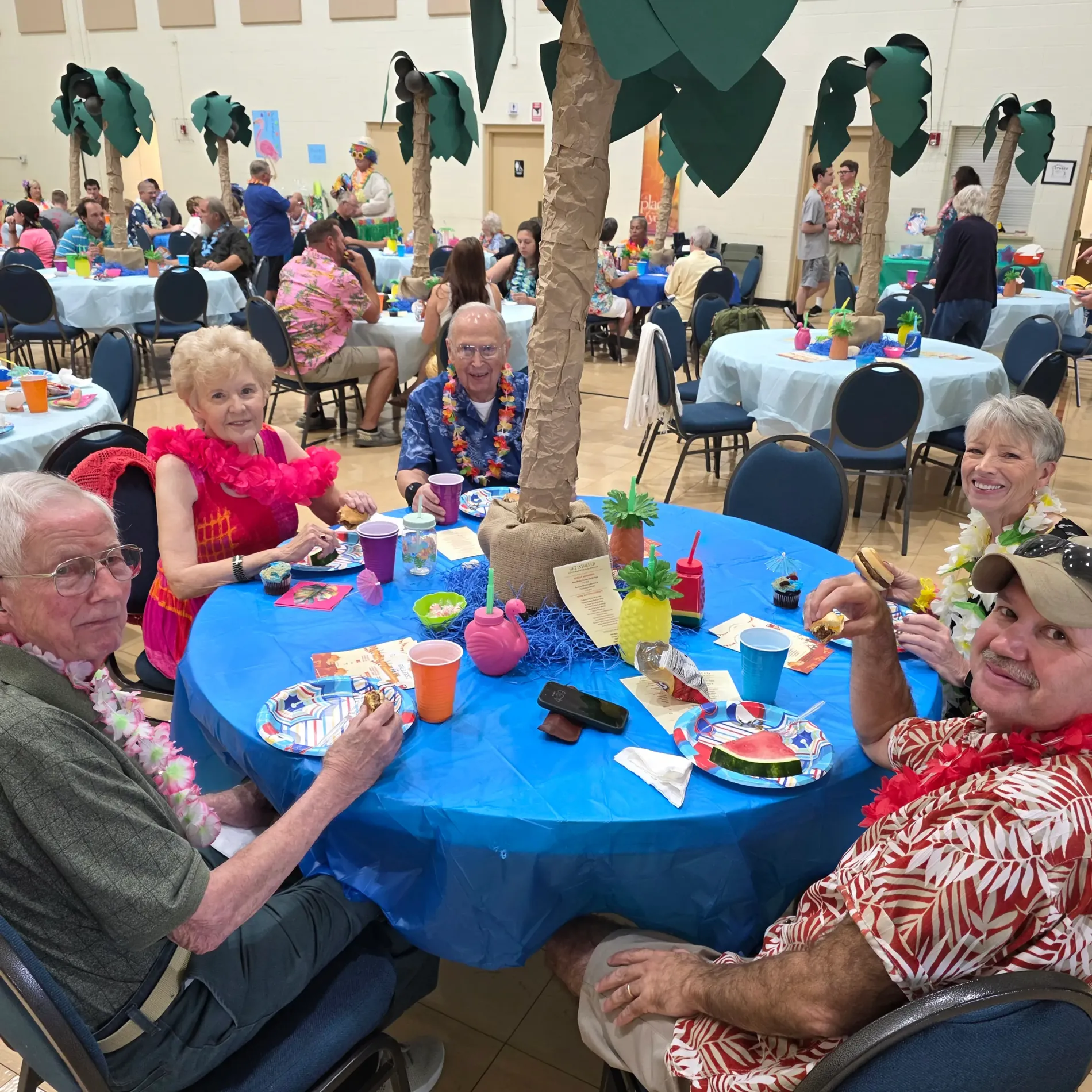 People enjoy a tropical-themed party, sitting around a table with palm tree decor.