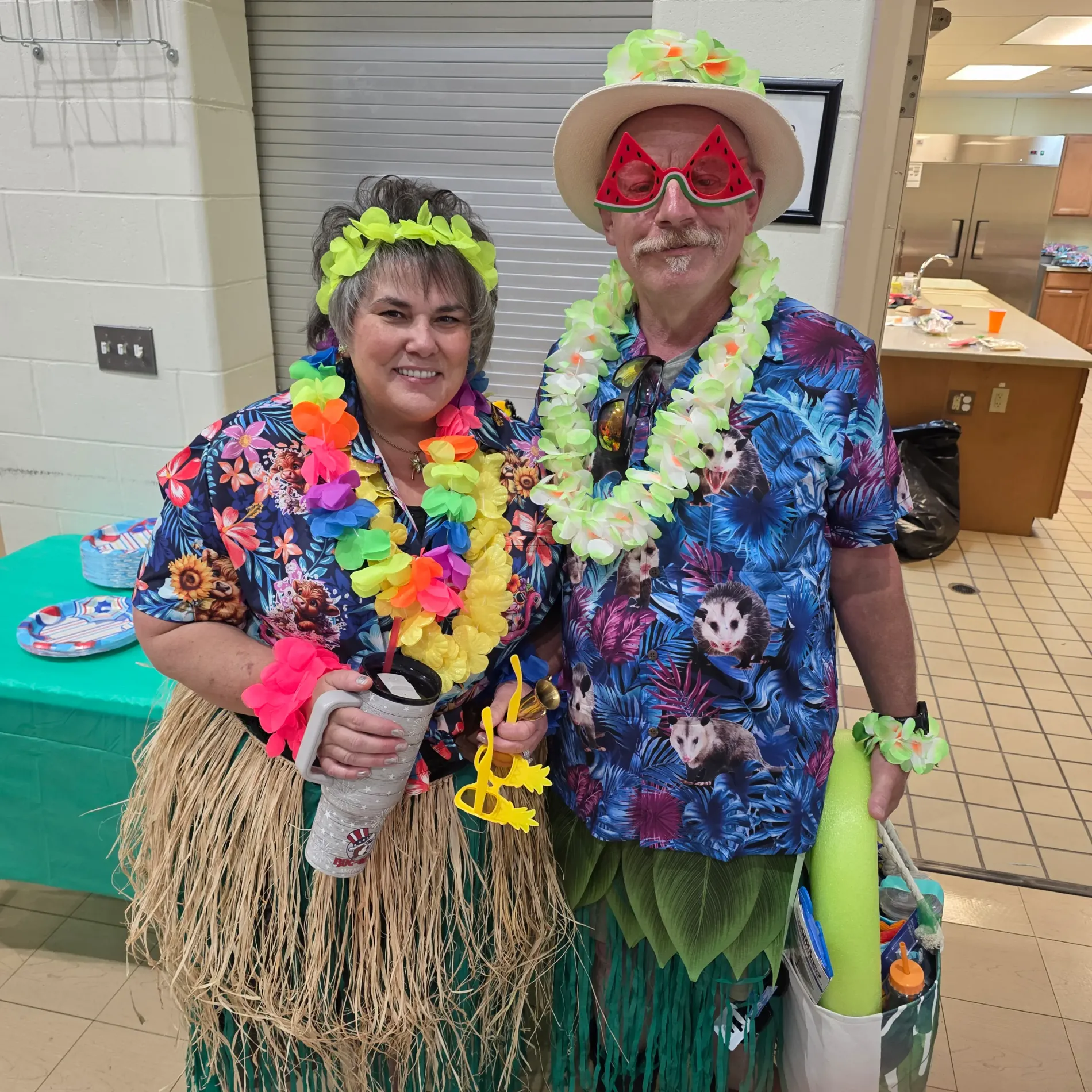 Two people in Hawaiian costumes with leis and grass skirts, smiling indoors.