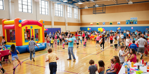 Busy indoor festival with children and adults, colorful bounce house, tables, balloons.